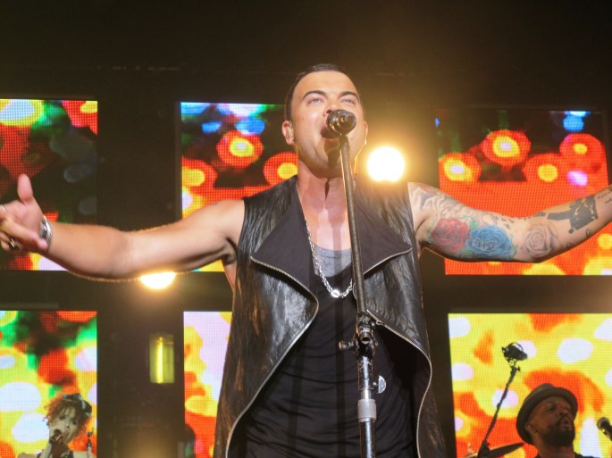 Guy Sebastian standing infront of a microphone singing with his arms outstreatched. He is wearing a black singlet and a grey singlet, with a black leather sleeveless jacket that is unzipped.
